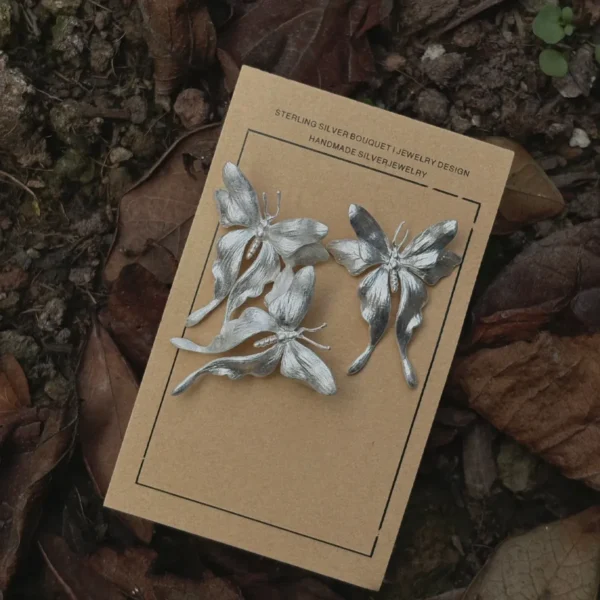 Asymmetric Butterfly Earrings in S990 Sterling Silver 05 Three handcrafted Butterfly Earrings in S990 sterling silver displayed on a card placed among dried leaves and soil, featuring sculpted wings and detailed textures in a nature-inspired setting.