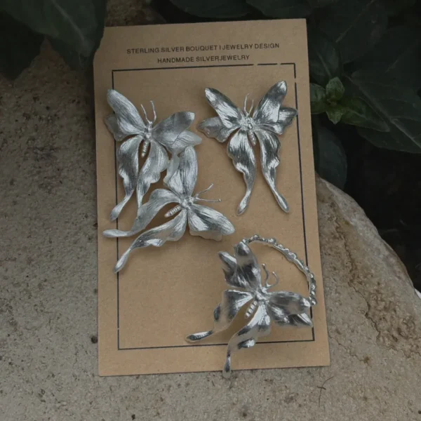 Asymmetric Butterfly Earrings and Butterfly Ring in S990 Sterling Silver 01 One handcrafted Butterfly Earrings and a Butterfly Ring in S990 sterling silver displayed on a brown card with green leaves, featuring sculpted wings and detailed textures in a natural setting.
