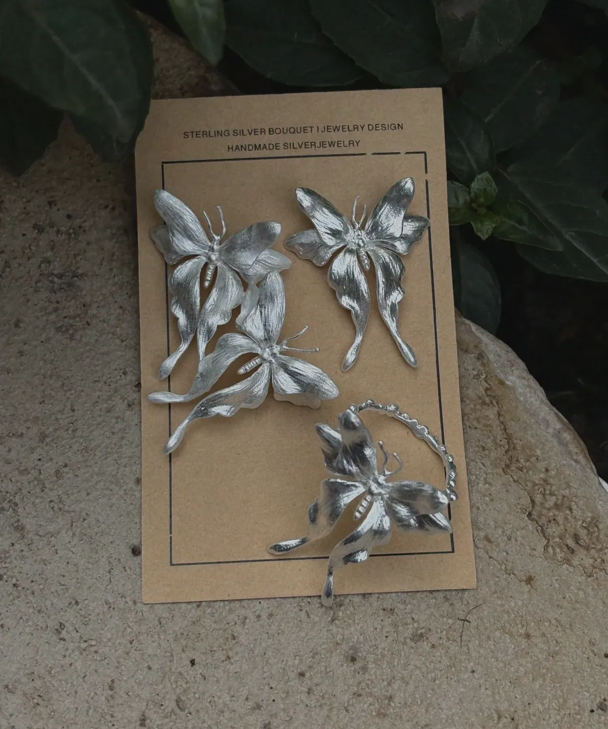 Asymmetric Butterfly Earrings and Butterfly Ring in S990 Sterling Silver 01 One handcrafted Butterfly Earrings and a Butterfly Ring in S990 sterling silver displayed on a brown card with green leaves, featuring sculpted wings and detailed textures in a natural setting.