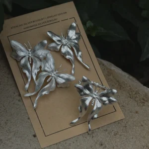 Handcrafted Butterfly Earrings and a Butterfly Ring in S990 sterling silver displayed on a brown card with floral and butterfly motifs, surrounded by natural greenery in a textured outdoor setting.