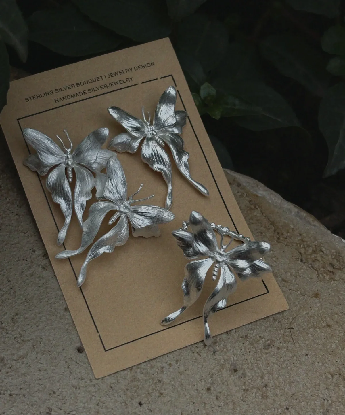 Asymmetric Butterfly Earrings and Butterfly Ring in S990 Sterling Silver 02 Handcrafted Butterfly Earrings and a Butterfly Ring in S990 sterling silver displayed on a brown card with floral and butterfly motifs, surrounded by natural greenery in a textured outdoor setting.
