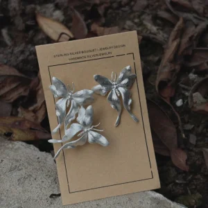 Asymmetric Butterfly Earrings in S990 Sterling Silver 08 Handcrafted Butterfly Earrings in S990 sterling silver displayed on a brown card set on natural soil and leaves, featuring sculpted wings and detailed textures in an organic outdoor setting.