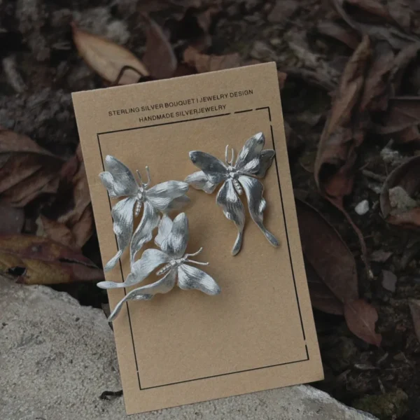 Asymmetric Butterfly Earrings in S990 Sterling Silver 08 Handcrafted Butterfly Earrings in S990 sterling silver displayed on a brown card set on natural soil and leaves, featuring sculpted wings and detailed textures in an organic outdoor setting.