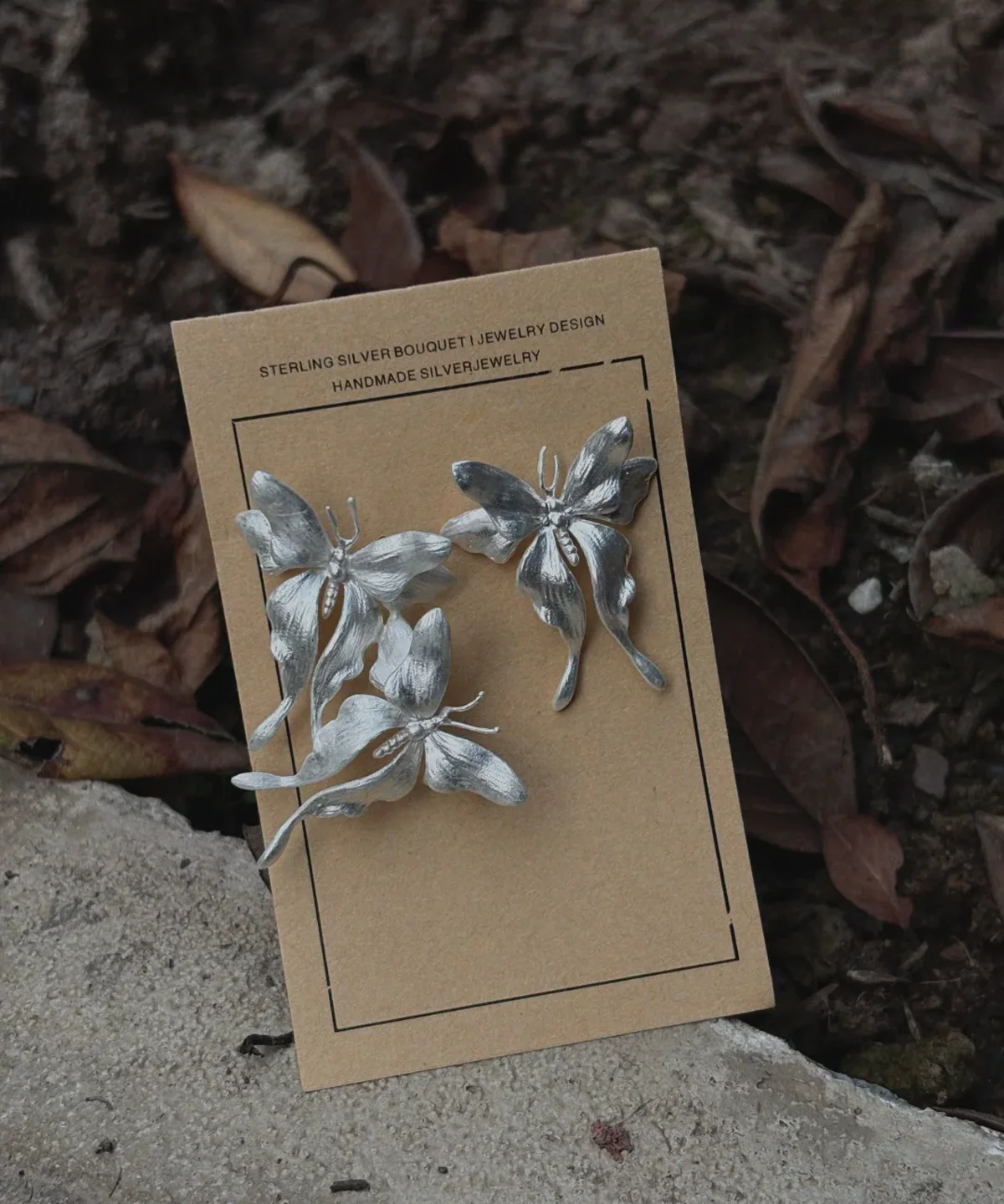 Asymmetric Butterfly Earrings in S990 Sterling Silver 08 Handcrafted Butterfly Earrings in S990 sterling silver displayed on a brown card set on natural soil and leaves, featuring sculpted wings and detailed textures in an organic outdoor setting.