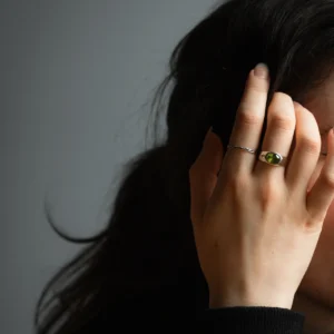 Hand wearing a silver August Birthstone Ring with a vivid green gemstone, partially covering the face, highlighting the Natural Peridot Ring against dark hair and a neutral background.
