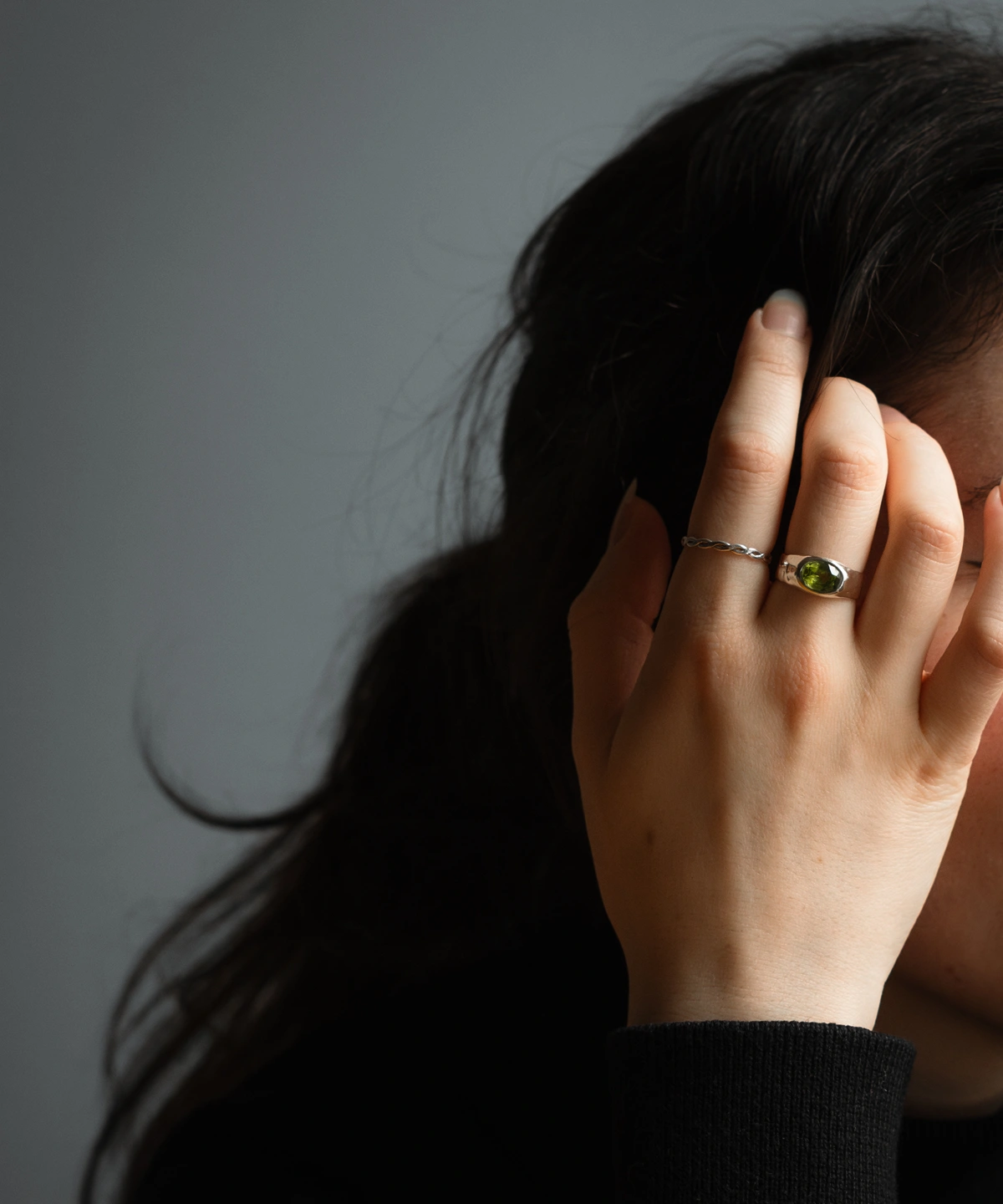 Hand wearing a silver August Birthstone Ring with a vivid green gemstone, partially covering the face, highlighting the Natural Peridot Ring against dark hair and a neutral background.
