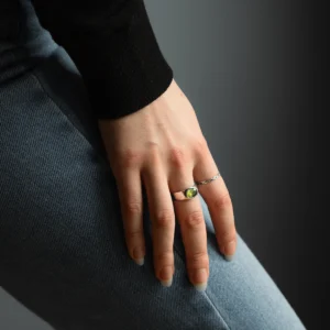 Hand wearing a silver August Birthstone Ring with a vivid green gemstone, resting on denim and a black sleeve, highlighting the Natural Peridot Ring design.