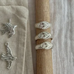 Hand wearing multiple rings, including a gold toned Bird Ring and a silver Signet Pinky Ring with raised hand‑shaped motifs, displayed on textured fabric.