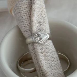 Small white bowl holding silver rings, including a Signet Pinky Ring with embossed clasping hands, displayed with rolled linen on a light fabric surface.