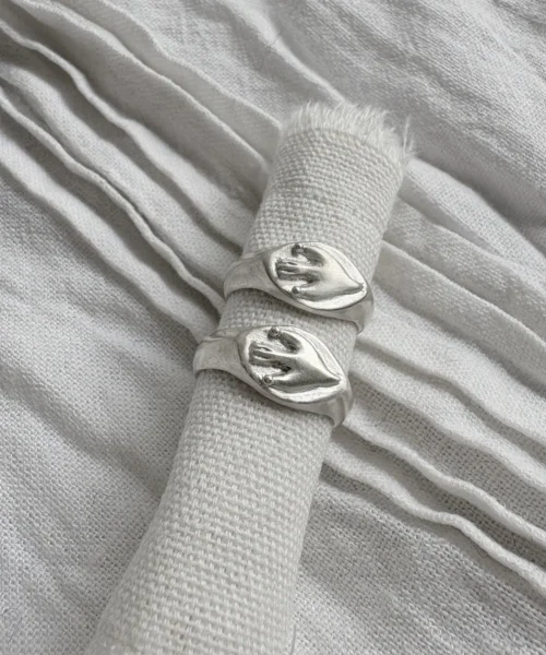 Two silver Signet Pinky Rings with raised hand motifs displayed vertically on rolled light fabric, surrounded by layered pleated textiles.