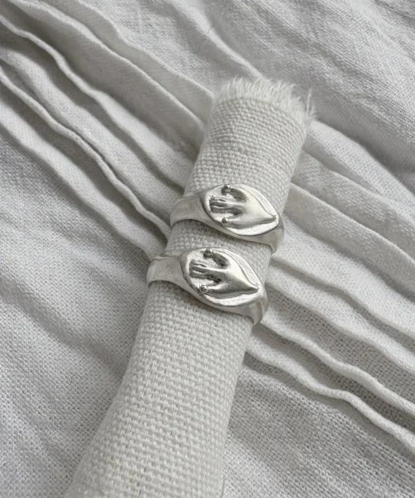 New In 23 Two silver Signet Pinky Rings with raised hand motifs displayed vertically on rolled light fabric, surrounded by layered pleated textiles.