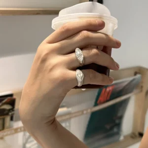 Hand holding a drink while wearing a silver Signet Pinky Ring with a sculpted face design, shown against a wooden shelf with magazines.