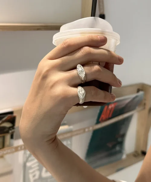 Hand holding a drink while wearing a silver Signet Pinky Ring with a sculpted face design, shown against a wooden shelf with magazines.