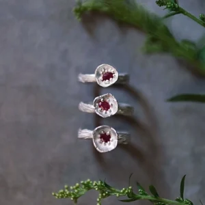 Three handcrafted S925 sterling silver rings with red gemstones arranged vertically on a gray surface with green sprigs, styled to highlight the artisanal design of a Ruby Ring product.
