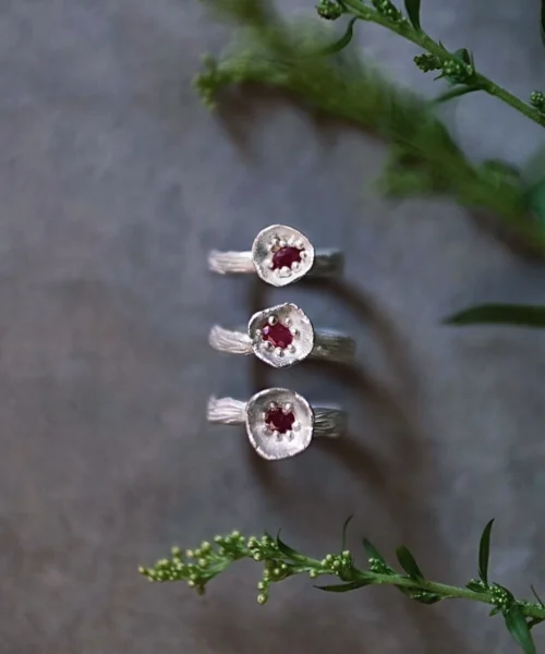 Three handcrafted S925 sterling silver rings with red gemstones arranged vertically on a gray surface with green sprigs, styled to highlight the artisanal design of a Ruby Ring product.