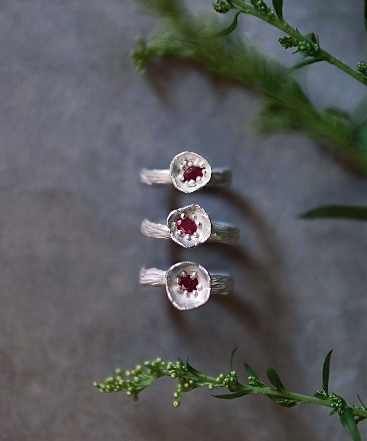 Three handcrafted S925 sterling silver rings with red gemstones arranged vertically on a gray surface with green sprigs, styled to highlight the artisanal design of a Ruby Ring product.