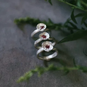 Poppy-Inspired Ruby Ring in S925 Sterling Silver 09 Three handcrafted S925 sterling silver rings with red gemstones displayed vertically on a dark surface with green foliage, supporting SEO for a Poppy‑Inspired Ruby Ring product.