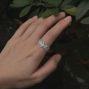 S925 Sterling Silver Poppy Leaf Ring Jewelry 01 Hand wearing a silver Leaf Ring Jewelry with delicate vine‑like leaf motifs, shown beside green foliage and a dark textured background.