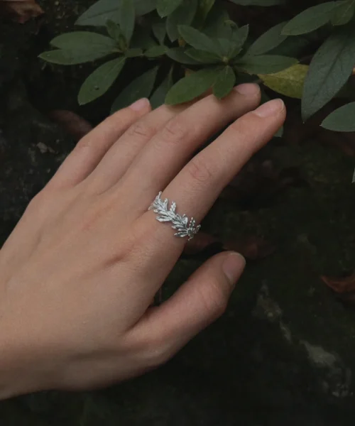 Hand wearing a silver Leaf Ring Jewelry with delicate vine‑like leaf motifs, shown beside green foliage and a dark textured background.