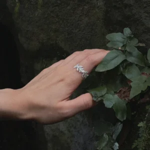 S925 Sterling Silver Poppy Leaf Ring Jewelry 02 Hand wearing a silver Leaf Ring Jewelry with delicate leaf motifs, touching green foliage on a mossy rock, highlighting its botanical and nature‑inspired design.