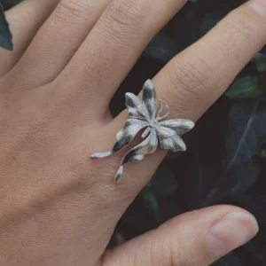 A handcrafted Sterling Silver Butterfly Ring in S990 silver worn on the finger, featuring sculpted petal-like details and a three‑dimensional organic form against a dark green foliage background.