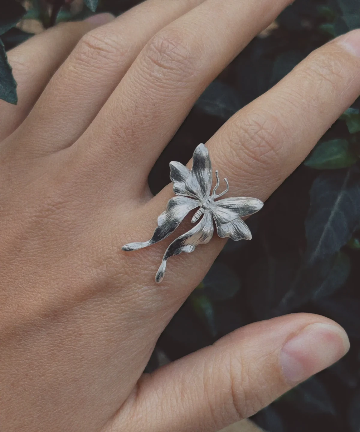 A handcrafted Sterling Silver Butterfly Ring in S990 silver worn on the finger, featuring sculpted petal-like details and a three‑dimensional organic form against a dark green foliage background.