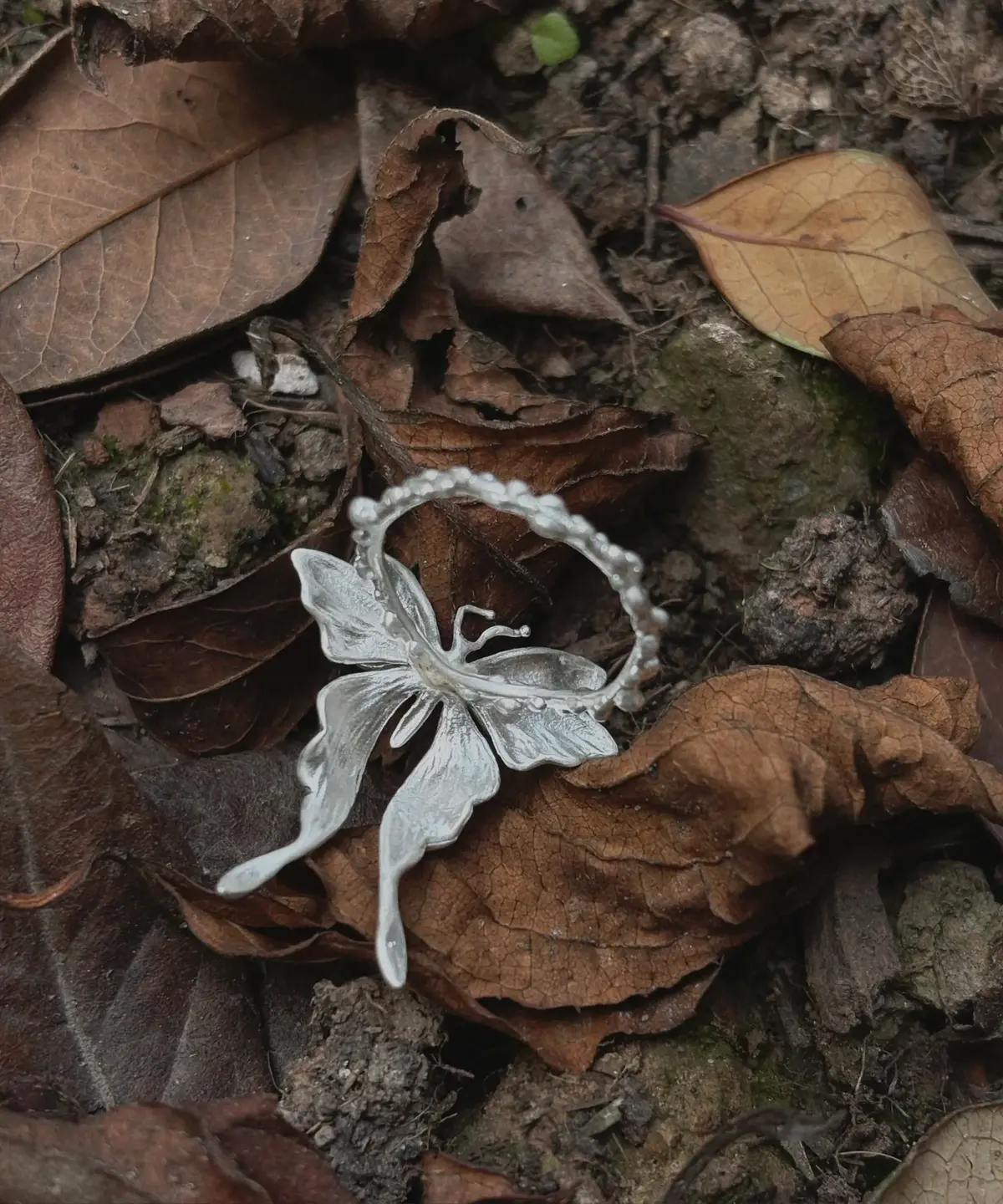 S990 Sterling Silver Butterfly Ring 02 A handcrafted S990 Sterling Silver Butterfly Ring resting on soil and dried leaves, featuring detailed wings and a textured band that highlights its organic, nature‑inspired design.