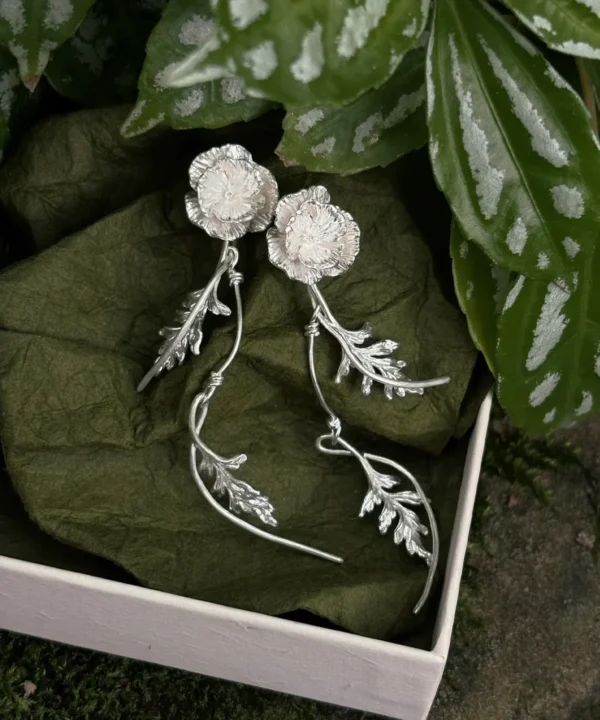 New In 14 A pair of elegant Poppy Earrings in S925 sterling silver, showcasing detailed poppy blooms and cascading leafy dangles, displayed in a gift box with natural greenery.