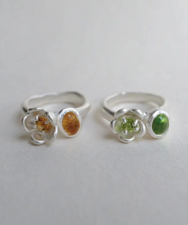 Two handcrafted silver peridot rings with double‑stone floral designs, featuring yellow‑orange and green gemstones placed on a light surface to highlight their polished finish and organic shapes.