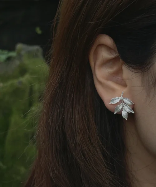 Close-up of a handcrafted S925 sterling silver Leaf Stud Earrings worn on an ear, showcasing delicate textured leaf details against a natural mossy background.