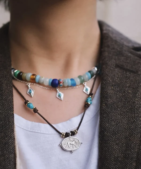 New In 11 A layered Horse Necklace featuring colorful beaded strands paired with a dark cord necklace and an engraved silver horse pendant, worn with a white shirt and brown textured outerwear.