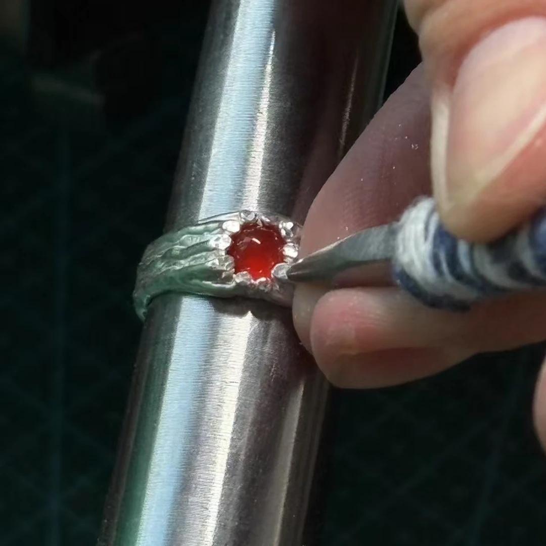 Hands adjusting silver around a garnet stone, showing the careful stone-setting process in artisan jewelry making.