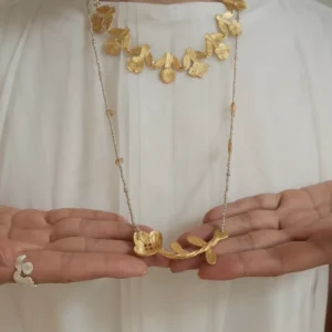 Short gold flower necklace worn at the collar, layered with a long beaded chain necklace held in open hands