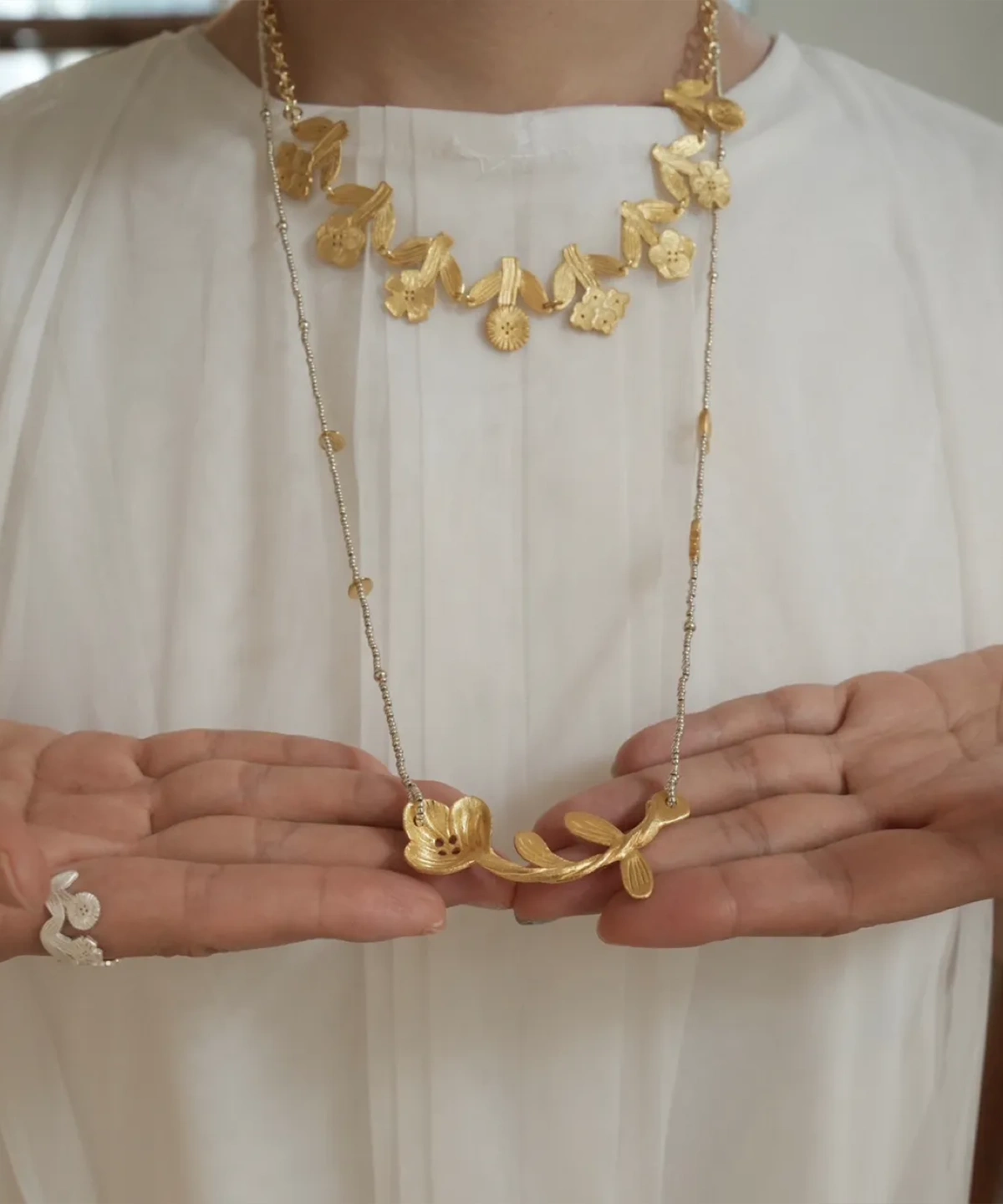 Short gold flower necklace worn at the collar, layered with a long beaded chain necklace held in open hands