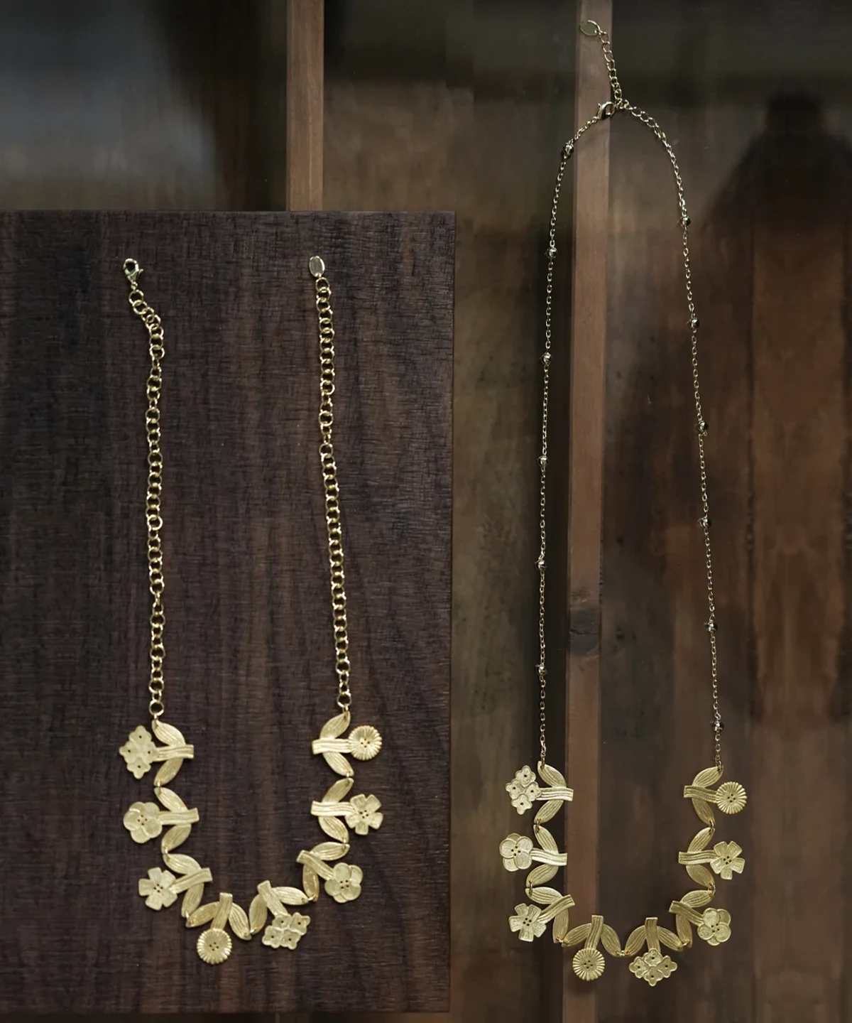 Gold flower necklaces in two lengths hanging on dark wood — short chain and long beaded chain, same linked garland