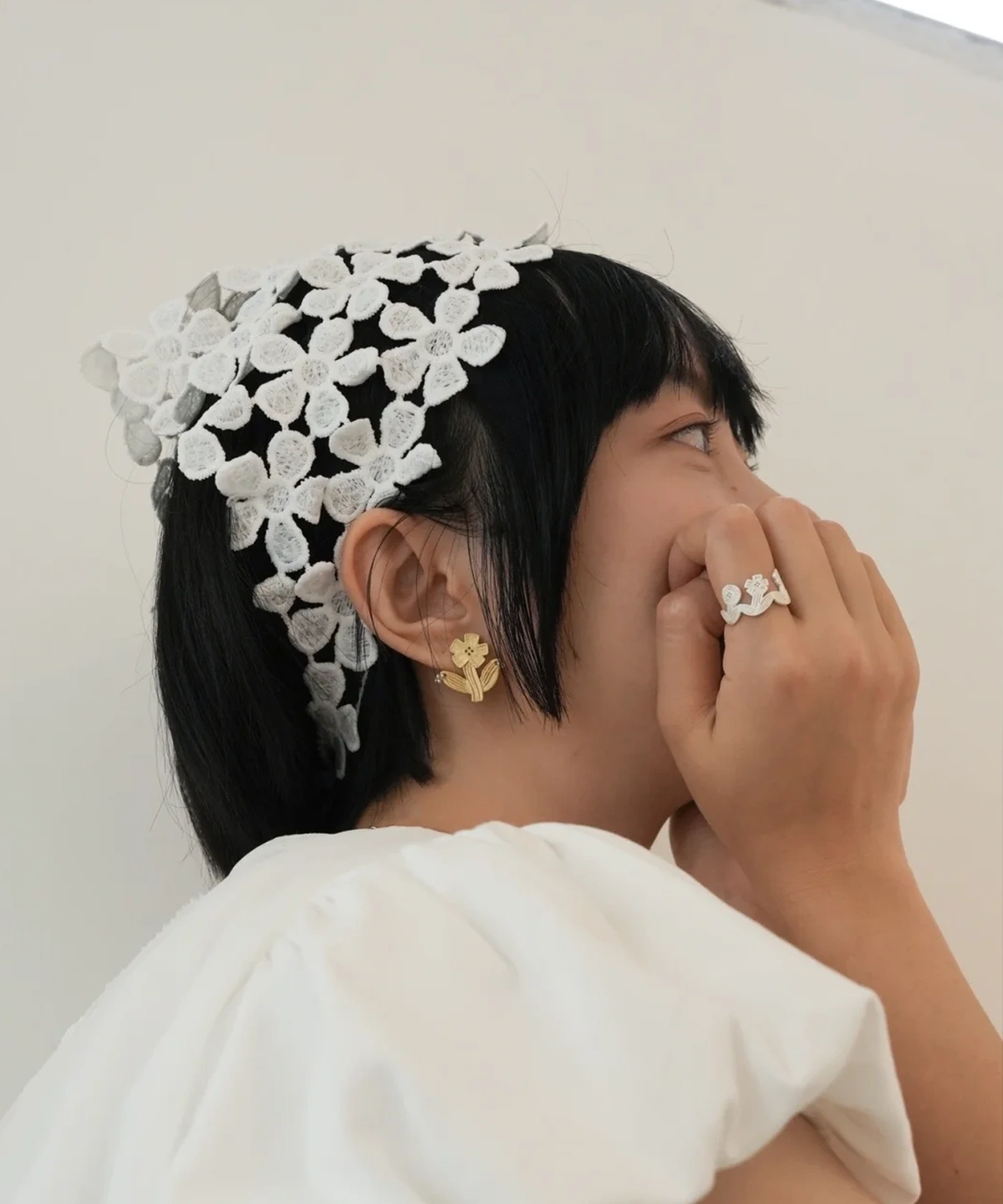 Gold flower stud earring worn with a white lace flower headband and a silver flower ring