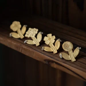 Four gold flower earrings in a row on a dark wooden shelf, whole flowers with stems and leaves