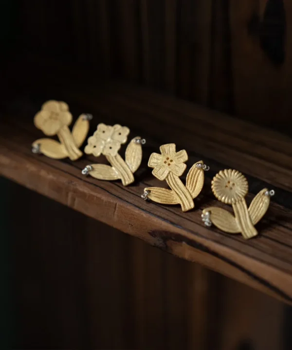 New In 3 Four gold flower earrings in a row on a dark wooden shelf, whole flowers with stems and leaves