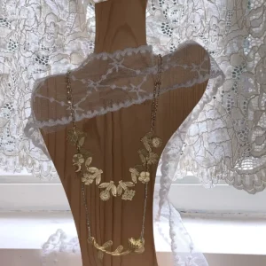 Two gold necklaces layered on a wooden display bust in front of a lace curtain