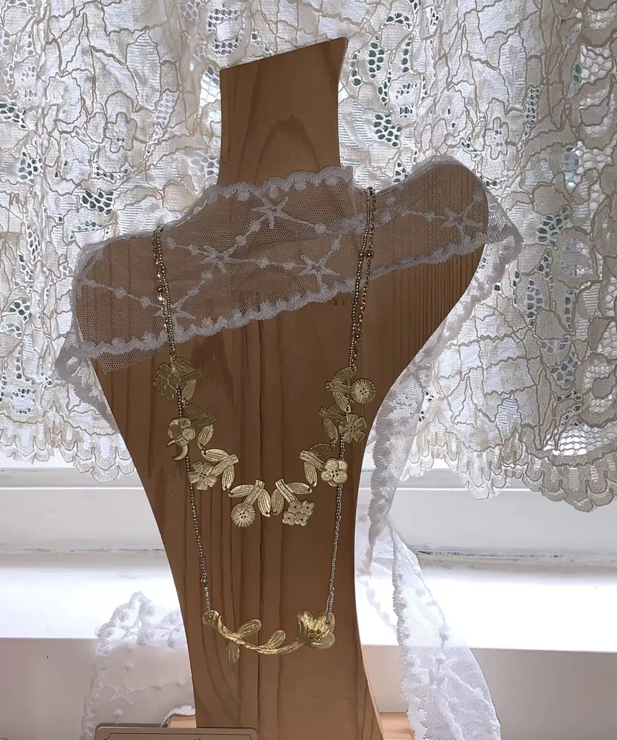 Two gold necklaces layered on a wooden display bust in front of a lace curtain