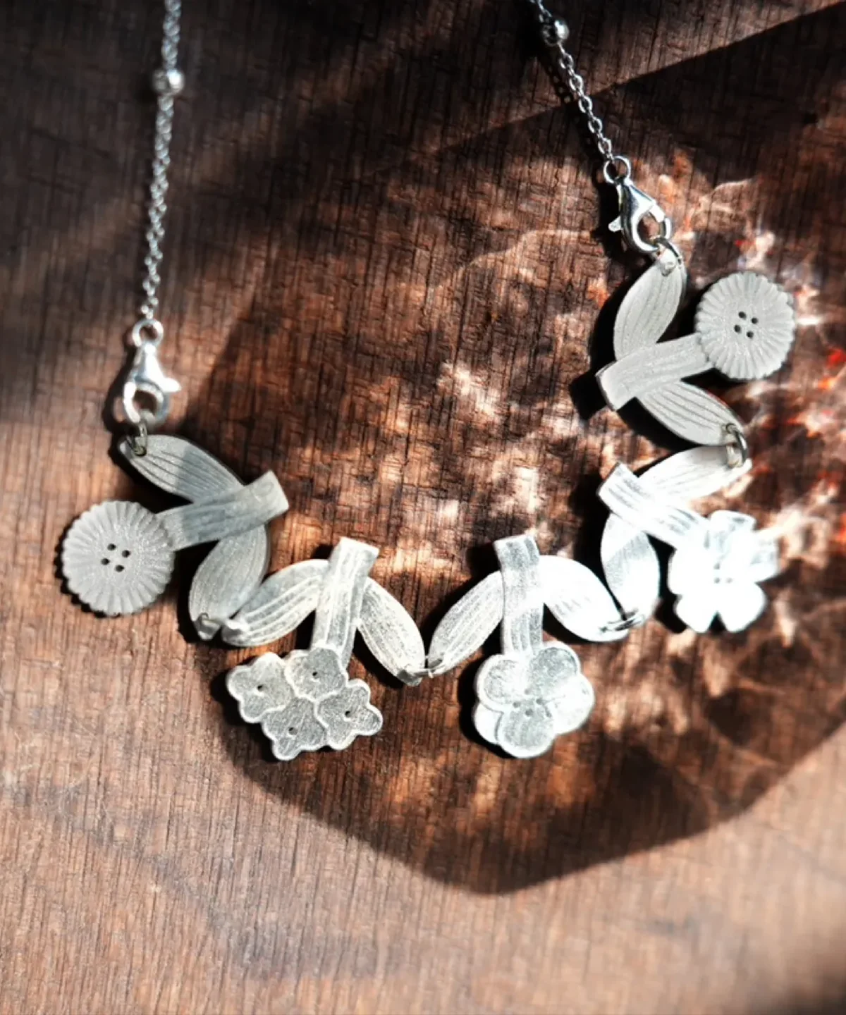 Silver flower necklace in dappled sunlight on dark wood — satin blooms, ribbed leaves, beaded chain