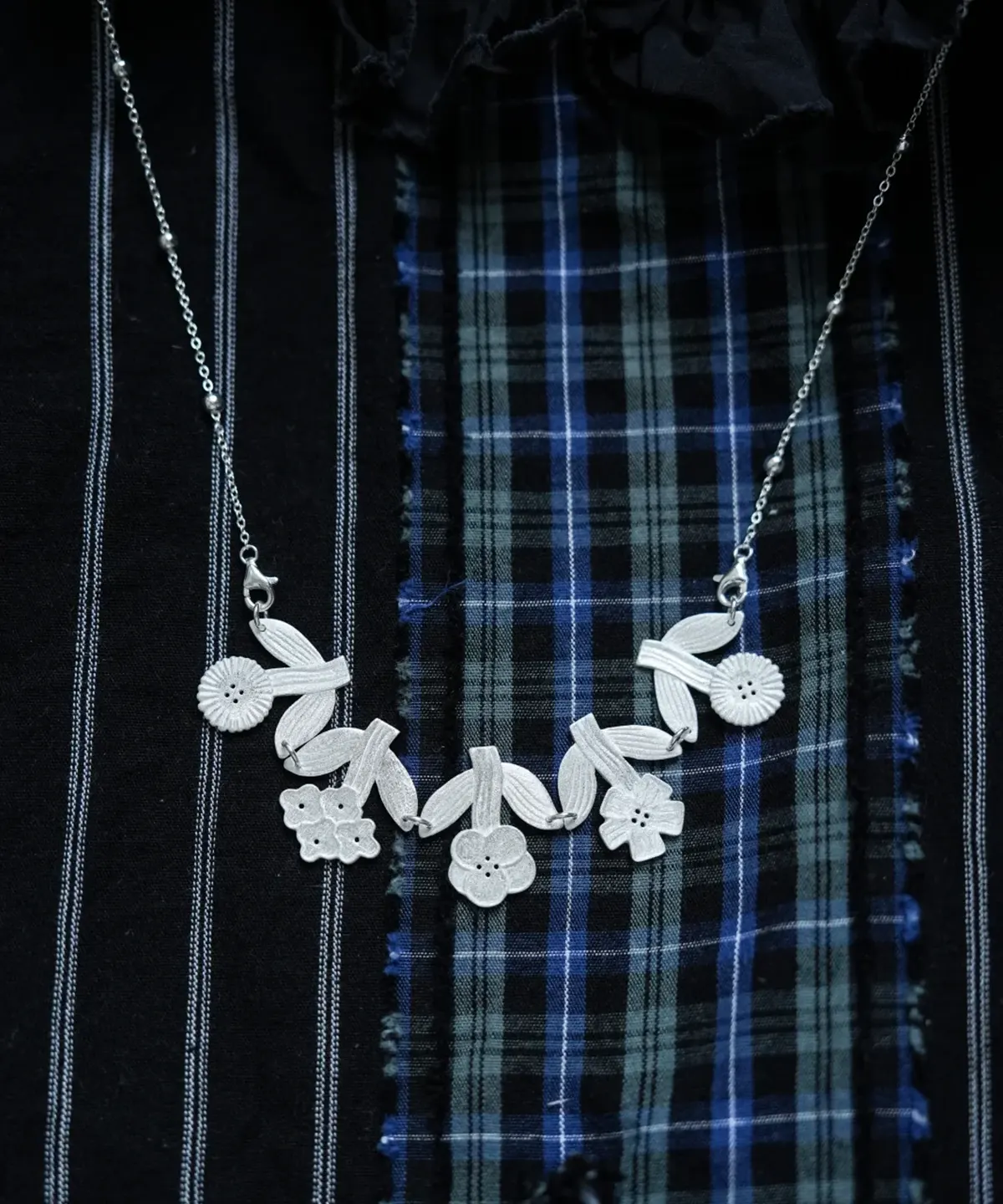 Silver flower necklace with linked blooms and leaves on dark plaid fabric, beaded chain