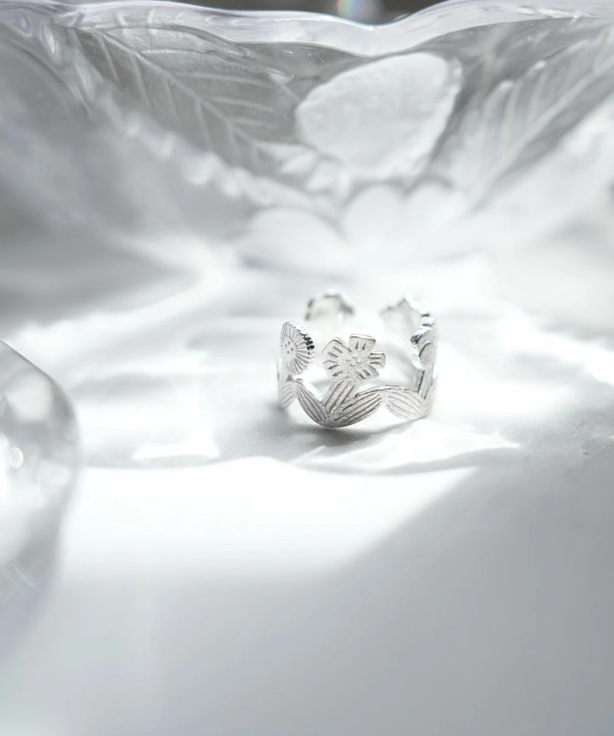 Silver flower ring standing on a white surface in rippled light, open garland band
