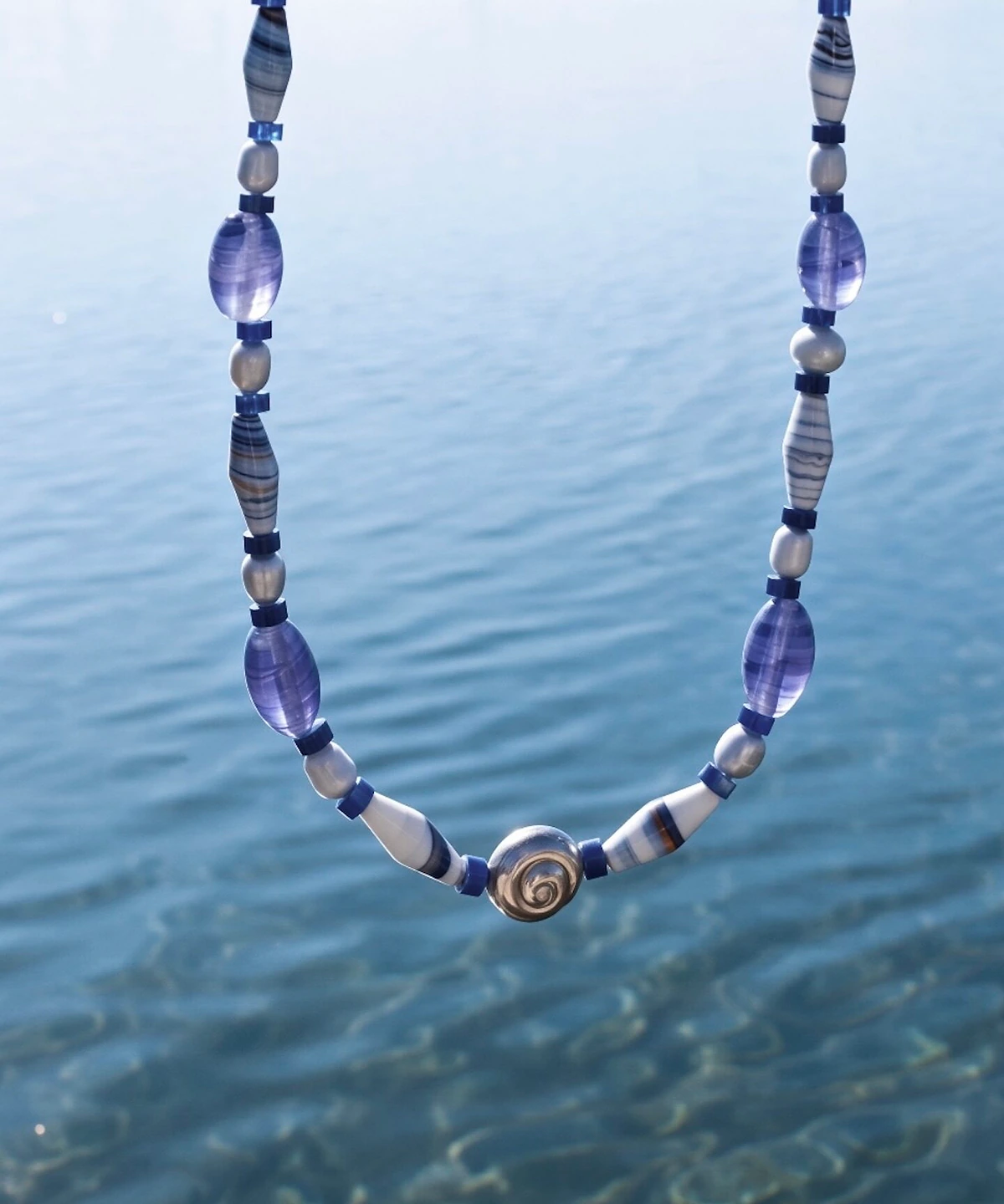 Blue agate necklace with a silver spiral shell centre held above clear sea water
