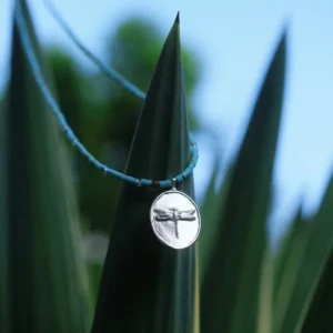 Silver dragonfly pendant necklace resting against long green leaves
