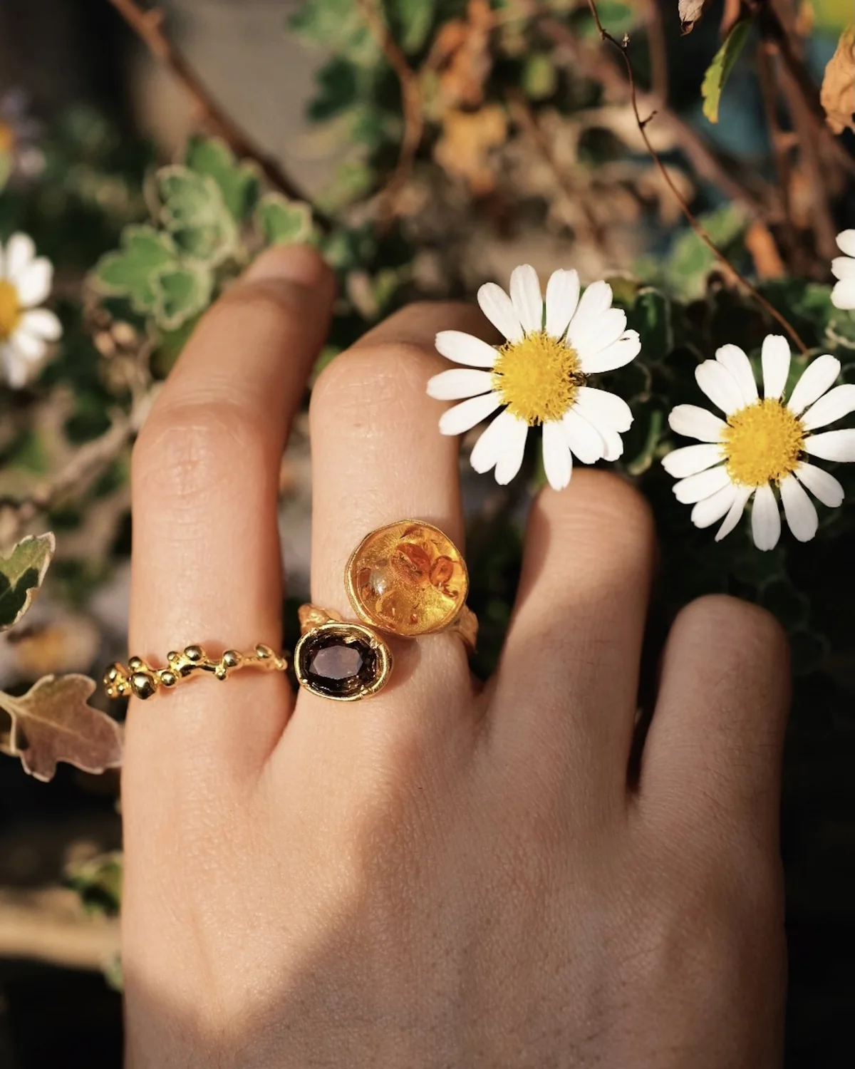 Warm gold rings with amber and dark stones layered beside small white flowers
