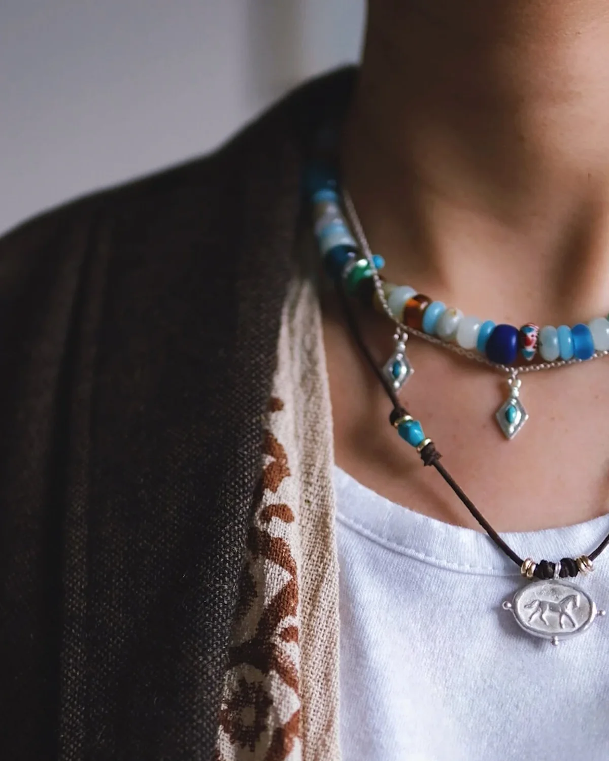 Mixed-Material Necklace Stack Detail Close detail of amazonite beads, turquoise silver charms and a horse pendant layered over a white top