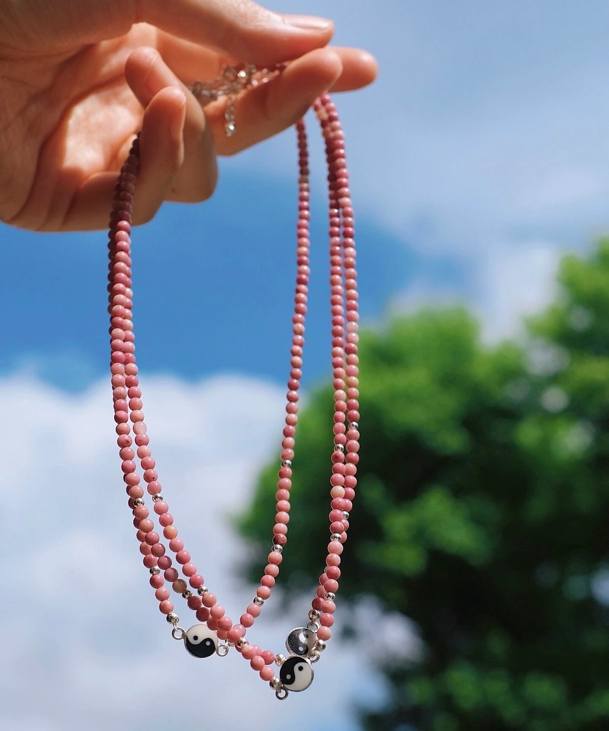 Rhodonite yin yang necklaces held in sunlight against sky and green leaves