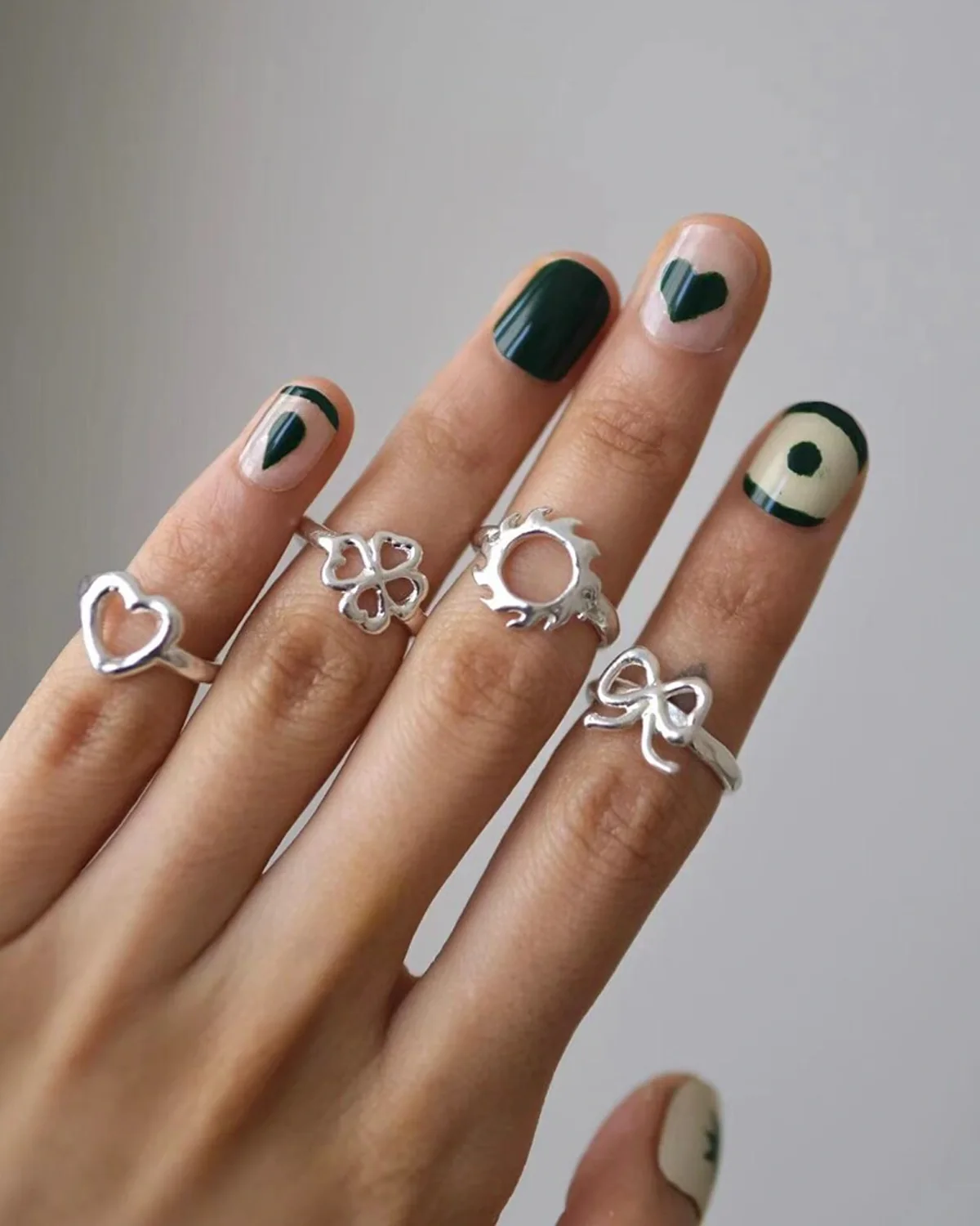 Thin silver stacking rings with heart, bow, clover, and sun symbols across the fingers