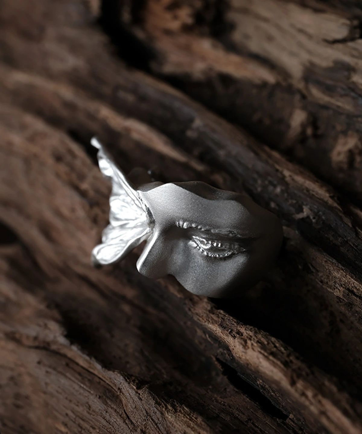 Close-up of the matte silver face on the Gaze ring, showing its sculpted eye and nose beside the wing
